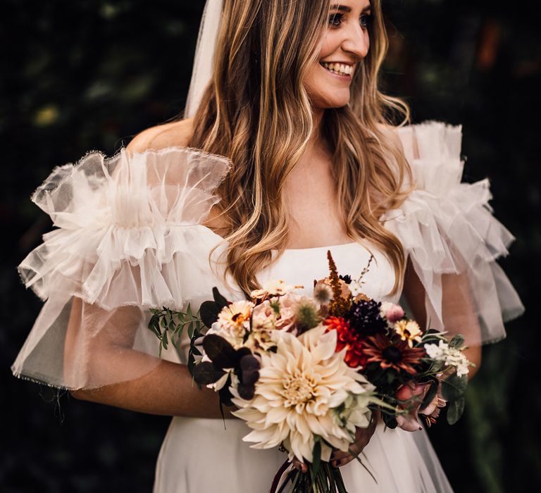 Bride in white Halfpenny London Mayfair dress with ruffle sleeves and veil stands holdingg mixed bridal bouquet with burgundy ribbon at garden wedding reception in Cornwall