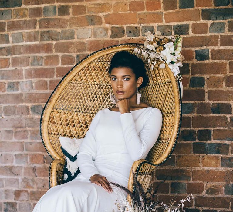 Boho bride in a fitted wedding dress sitting in a wicker peacock chair with dried flower and orchid wedding flower decor 