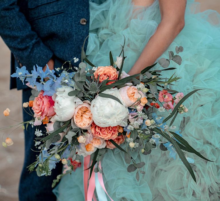 Bride in coloured tulle wedding dress holding a white, peach, pink and green wedding bouquet by her side