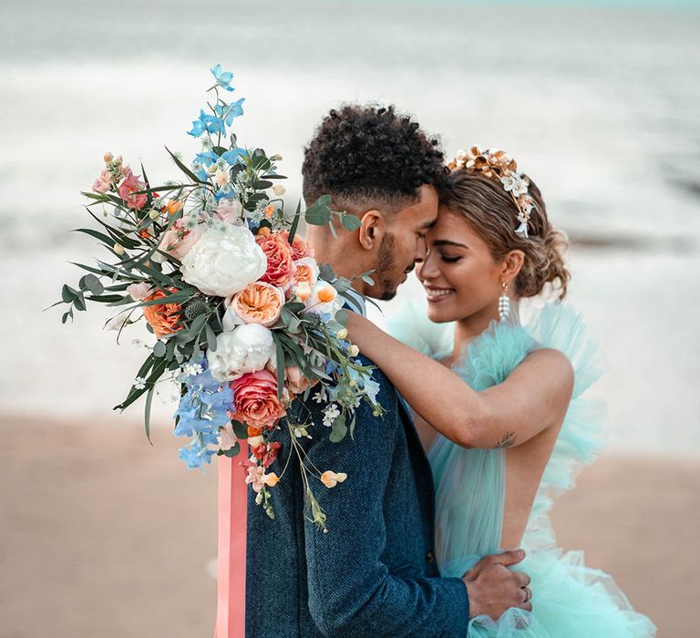 Intimate coastal wedding with bride in a mint green wedding dress holding a pastel flower wedding bouquet tied with ribbon over her grooms shoulder in a blue wool suit 