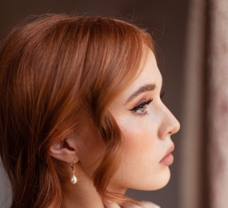 Bride with curled hair wearing pearl drop earrings and long sleeve wedding dress looks out of window