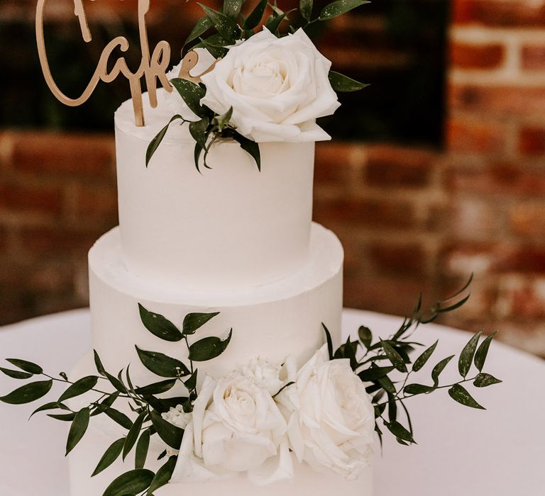 Elegant white buttercream wedding cake with foliage decor and gold lasercut cake topper sign 