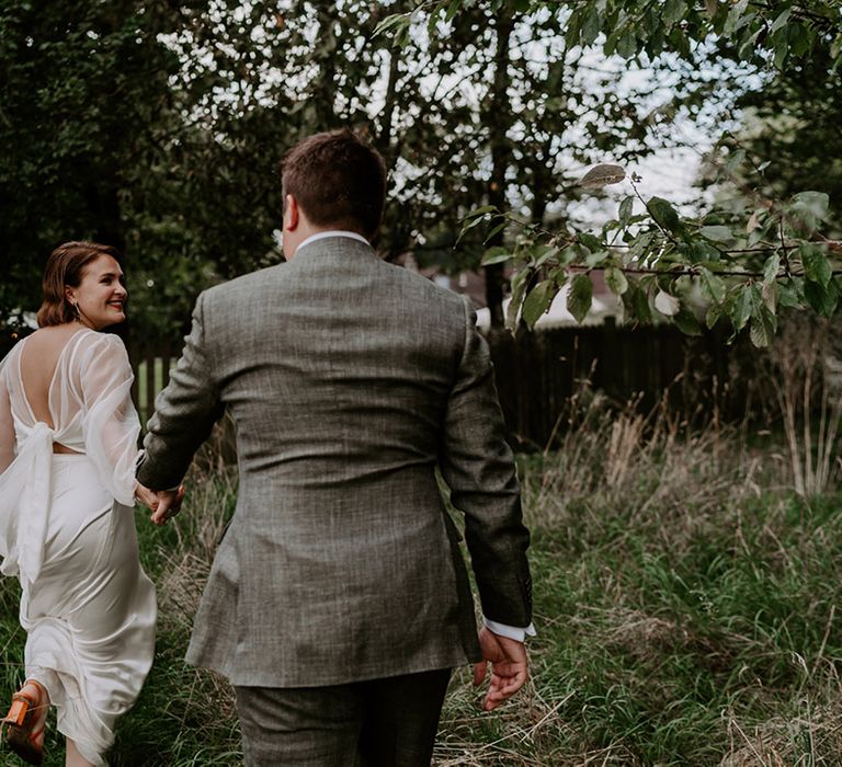 Groom in a grey check suit holding hands with his bride in a satin skirt and sheer blouse 