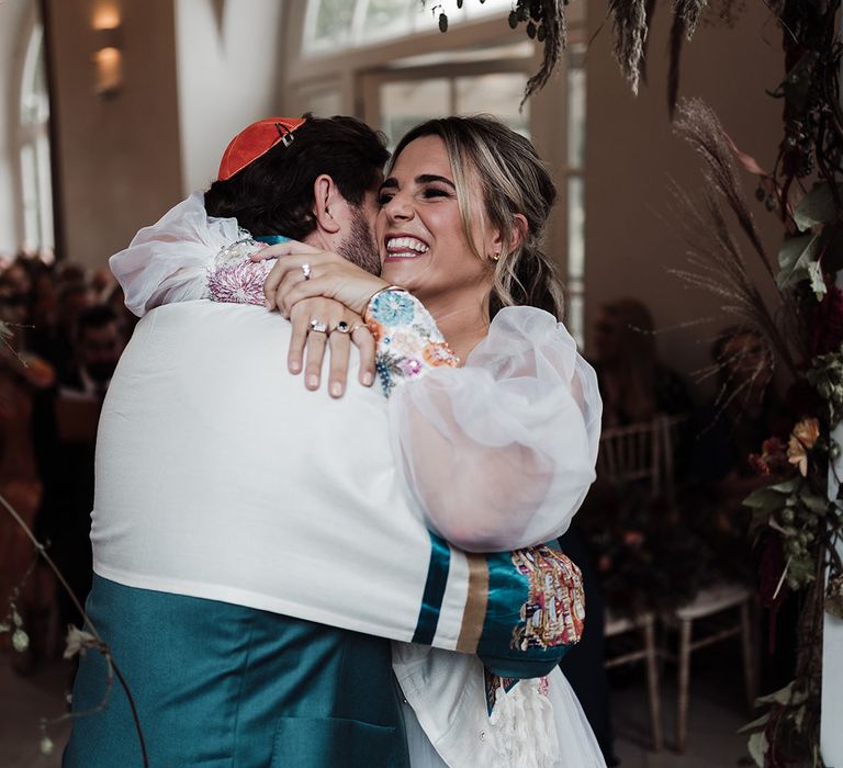 Bride in mesh sleeve wedding dress with embroidered floral cuffs hugs groom in red kippah, tallit and green suit during wedding ceremony