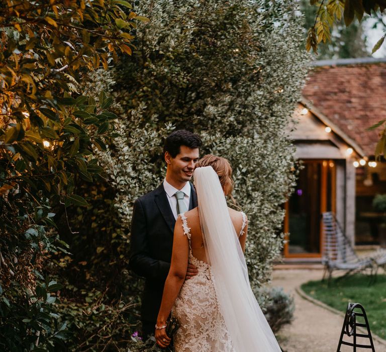 Bride in open back lace and tulle wedding dress with train and veil stands with groom in dark suit and sage green tie in the grounds of The Tythe Barn in Launton