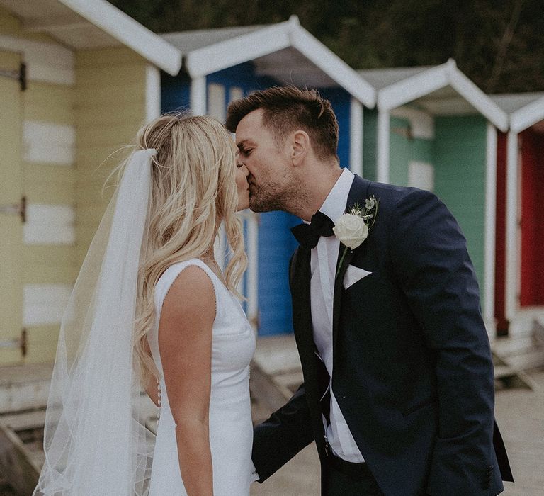 Bride holds large white flower bouquet as she kisses groom in classic black tuxedo