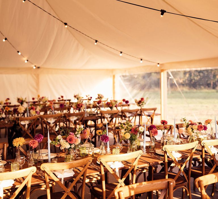 Rustic marquee wedding reception with long wooden tables and chairs, colourful florals and candles