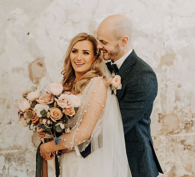 Bride in pearl wedding dress with mesh sleeves stands holding blush pink bridal bouquet with roses and anemones as groom in navy tweed suit hugs her