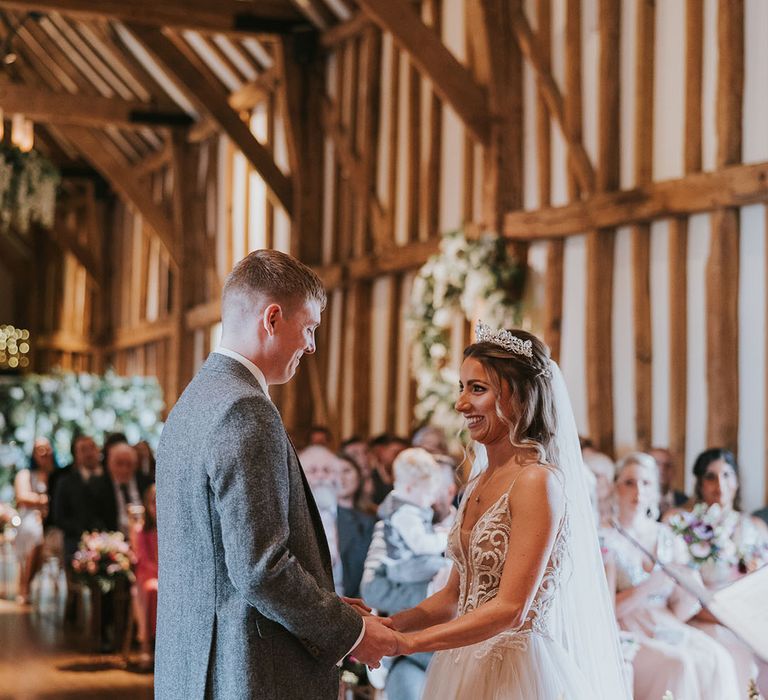 Bride in jewelled silver tiara and groom at the end of the aisle in rustic wedding venue complete with hanging wisteria 