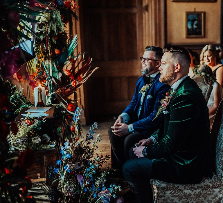 Grooms seated in damask pattern chairs for ceremony at Castle Leslie Estate with brightly coloured floral display