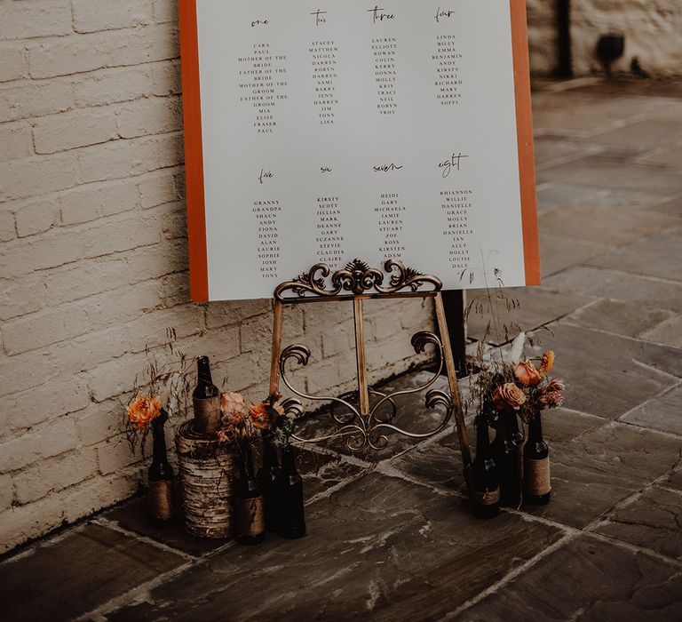 Orange and white seating chart sign for rustic wedding on gold stand with bottle vases and orange roses