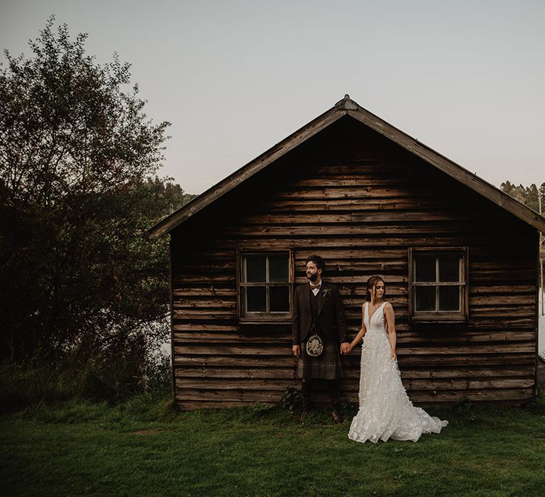 Groom in traditional kilt with bride in Savin Lond wedding dress hold hands in front of the boathouse at Cardney Estate wedding venue 