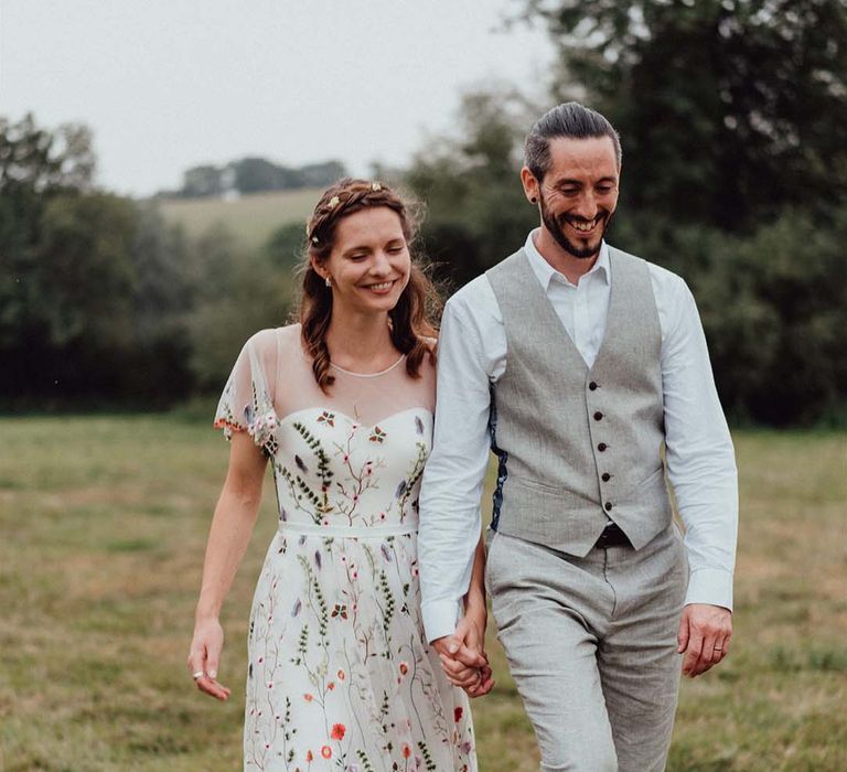 Bride in floral embroidery wedding dress with braided hair and flower accessories with groom in white shirt and grey trousers and waistcoat