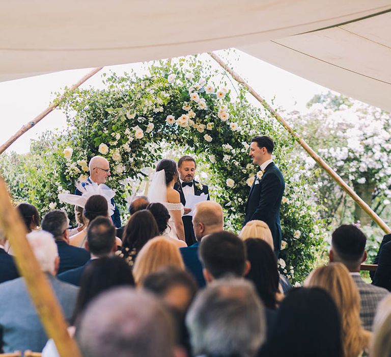 Bride and groom have outdoor wedding ceremony with large white florae and greenery arch at the altar 