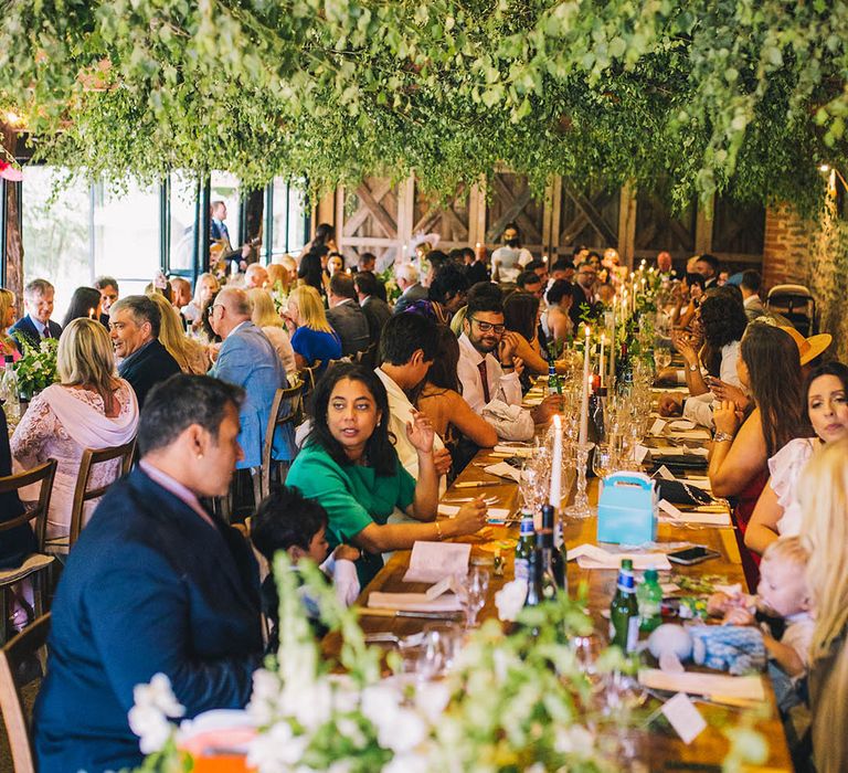 Wedding guests socialise while seated with greenery overhead 