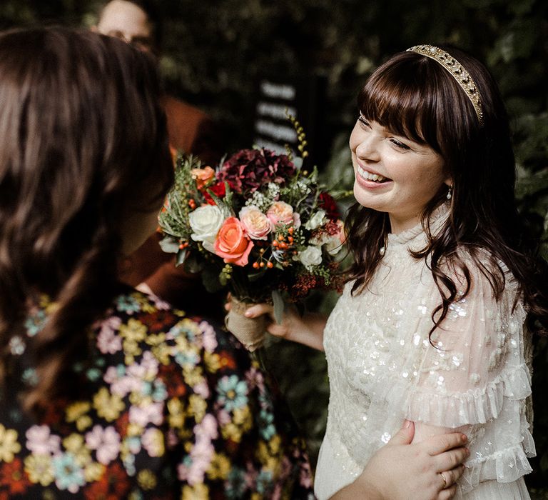 Bride socialises with guest wearing golden headband accessory, sparkly Needle and Thread top and orange, pink and red bouquet 