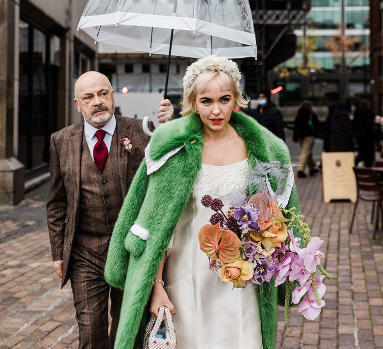 Father of the bride in checkered three piece suit holds an umbrella over the bride as she walks in her platform block heels and green fur coat