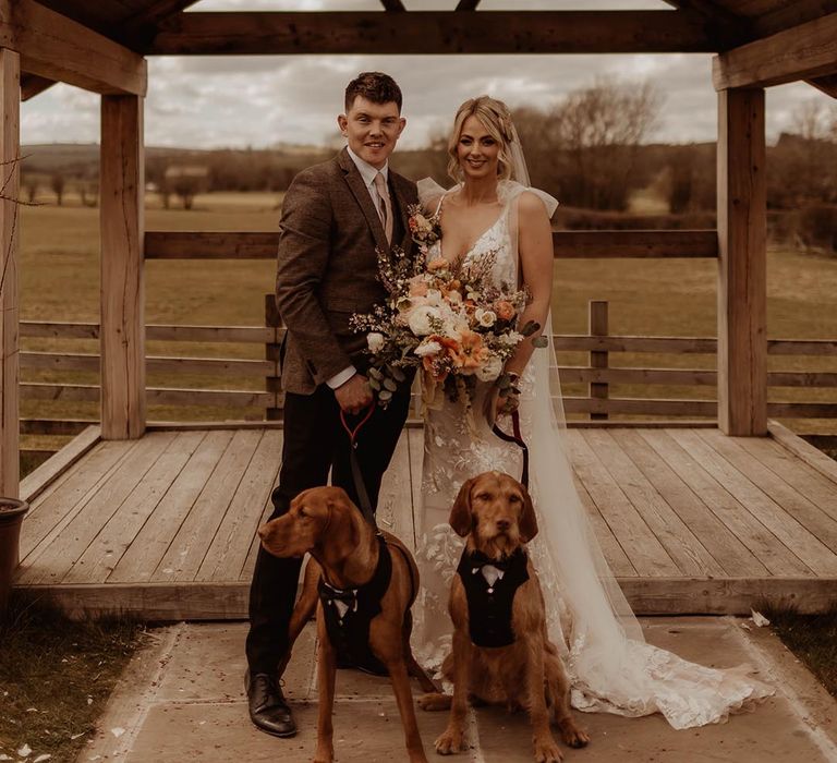 Rustic Eden Barn wedding with bride and groom holding their pet dogs on a lead wearing tuxedos 