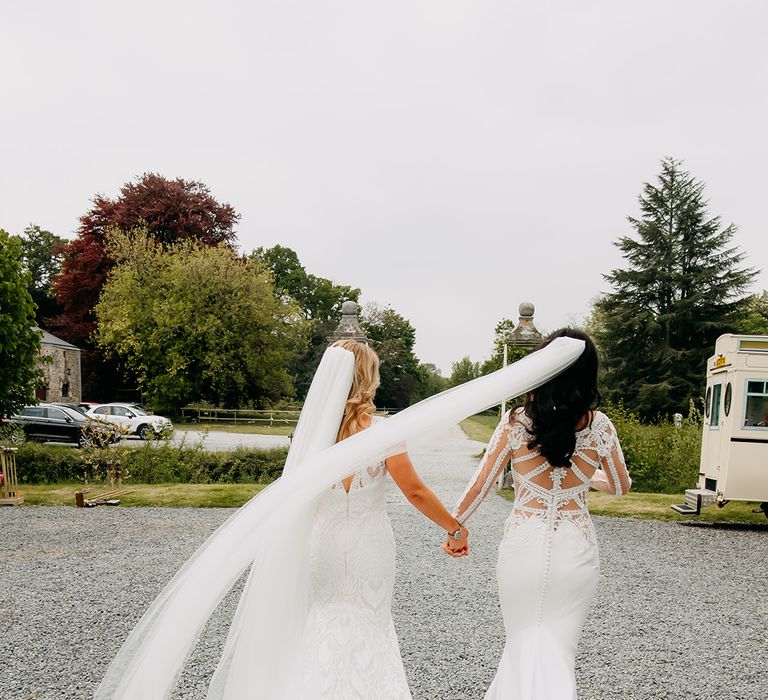 brides walk holding hands together with veils blowing in the breeze