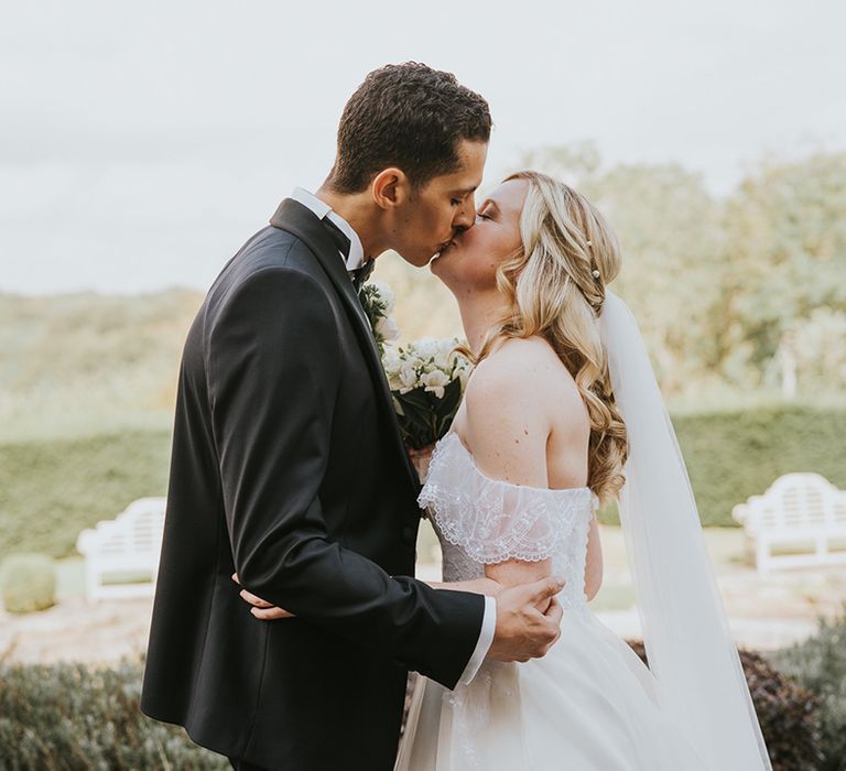 Bride in off the shoulder wedding dress with pearl hair accessory kisses groom in black tie