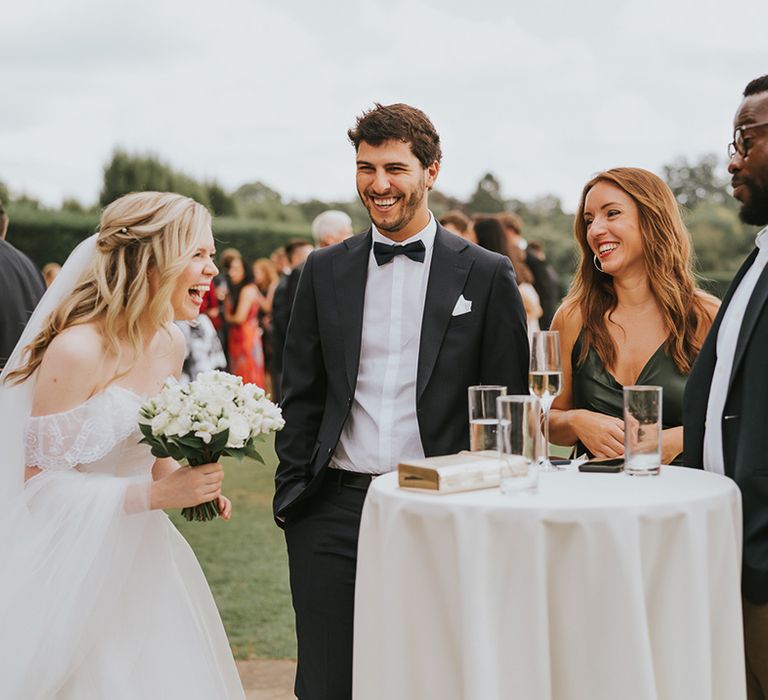 Bride holding white rose wedding bouquet socialises with guests outside around white circle tables 