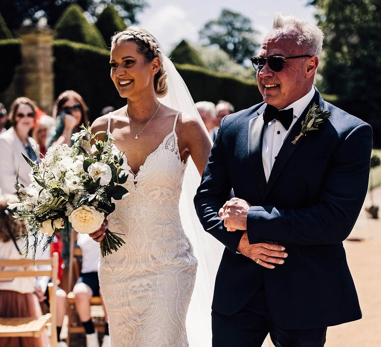 Father of the bride in sunglasses and black tie walks the bride in lace wedding dress down the aisle for outdoor wedding ceremony 