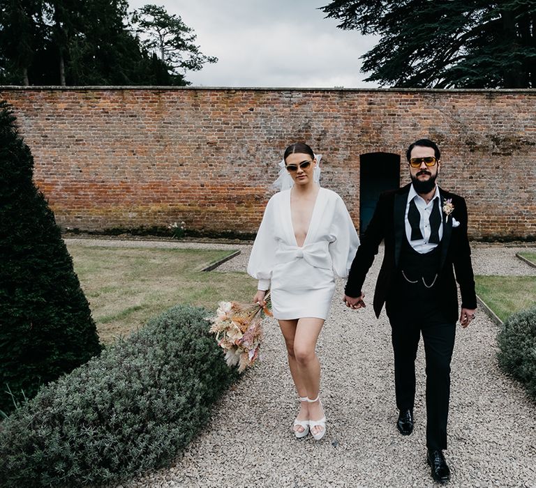 Groom in a horseshoe waistcoat with bow tie and chains, with his bride in a short wedding dress, sunglasses and platform shoes in Garthmyl Hall gardens  