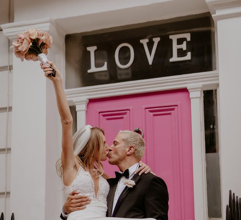 Groom in black tie carries the bride in front of bright pink door 