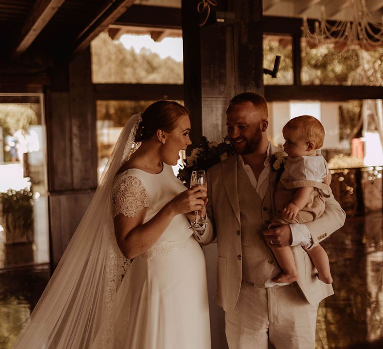 Bride holds glass of champagne as she stands beside her groom who carries their baby on the day of their wedding 