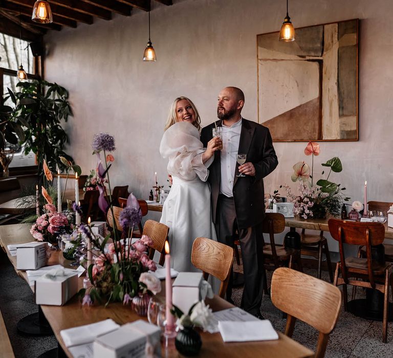 Bride and groom standing in the middle of their wedding reception with tall purple allium stems, pink anthuriums and taper candle table decor 