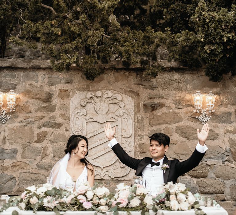 Bride & groom sit at sweetheart table finished with white linen tablecloth and floral bouquets to the front 