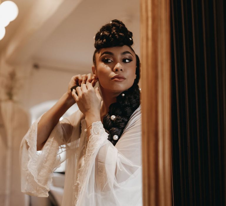 Bride wears sheer lace dressing gown and silk pyjamas whilst adding earrings on the morning of her wedding day