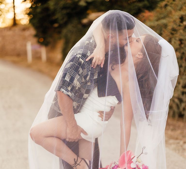 Bride wears white mini dress and holds brightly coloured bouquet as she kisses her groom beneath veil