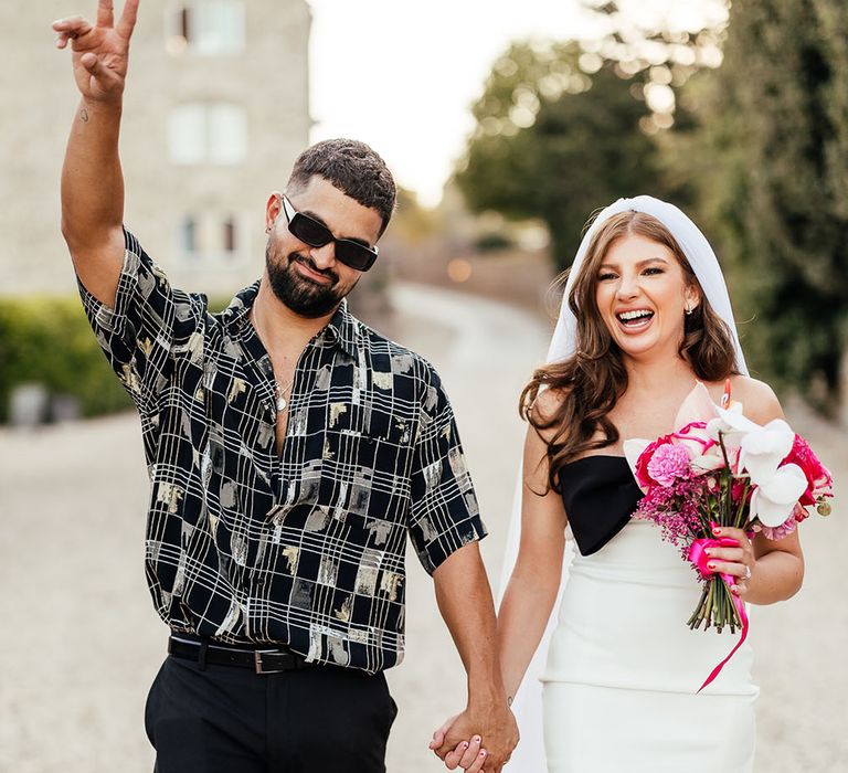 Groom wears open shirt and sunglasses whilst holding his brides hand who wears mini wedding dress 