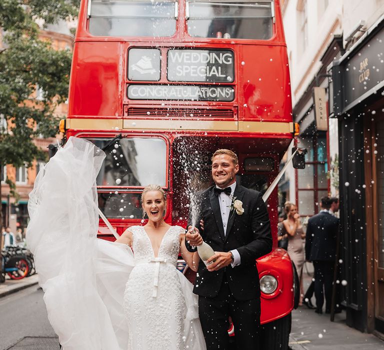 Groom wearing black tie pops bottle of champagne in front of red London bus whilst stood beside his bride in detachable skirt wedding dress