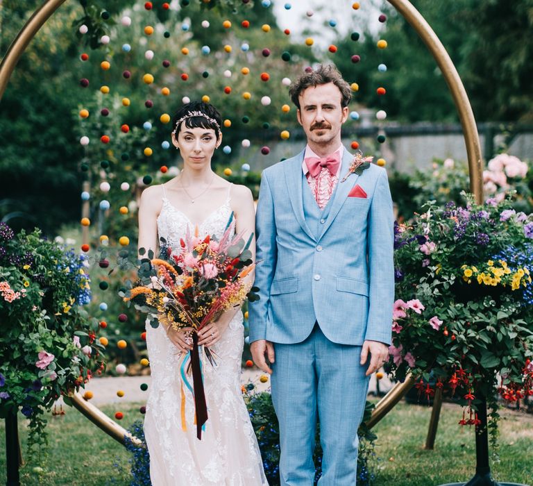 Bride wearing lace Enzoani wedding dress and groom in dusty blue suit stand in front of DIY colourful pom pom wedding decorations 