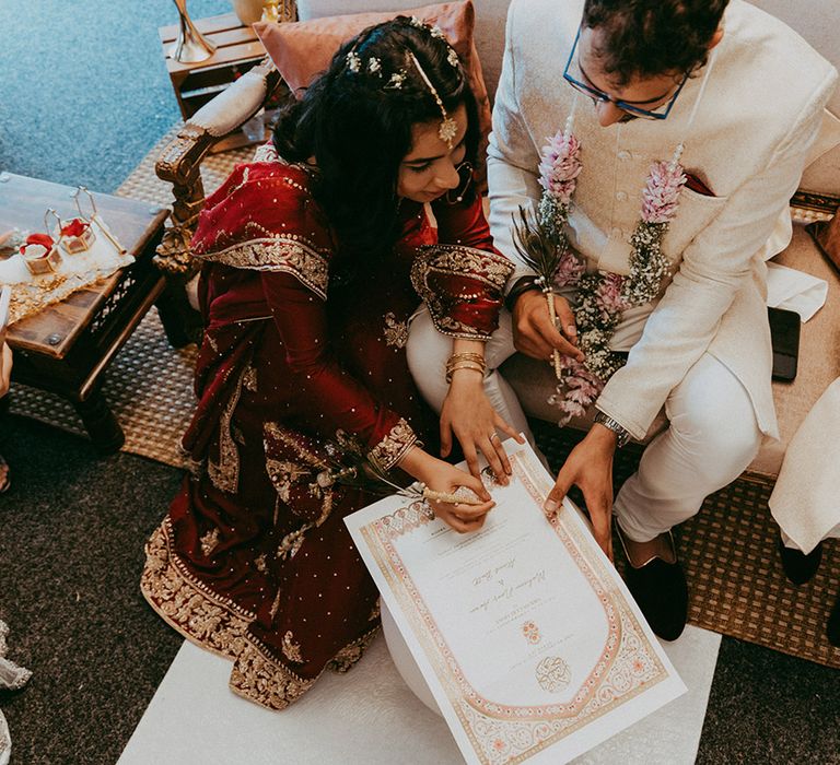 Bride wears gold hair accessories and sits beside her groom as they sign marriage certificate during traditional Nikkah ceremony