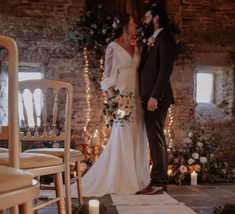 Bride and groom stand at the altar with pillar candles, white and blush pink roses with foliage and fairy lights 