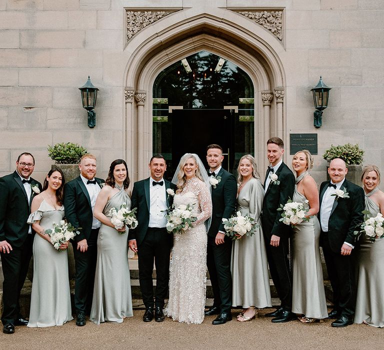 Bride & groom stand with their wedding party with bridesmaids in sage green bridesmaid dresses and groomsmen in black tie 