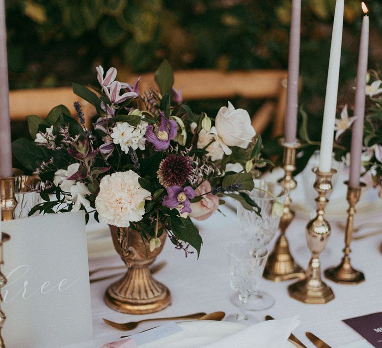 Floral arrangement with white carnations, purple tulips, roses and foliage in gold vase, white and lavender tapered candles in gold candle holders, and gold cutlery