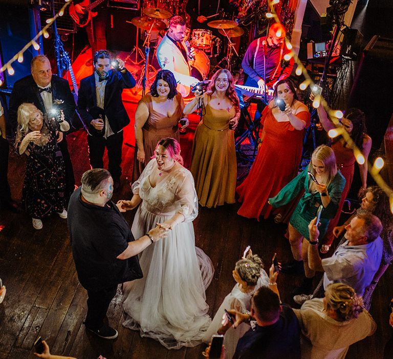 Bride in corset top wedding dress with leaf detailed lace overlay, pink stitching around the waist and red gemstone necklace dancing with groom in black shirt 