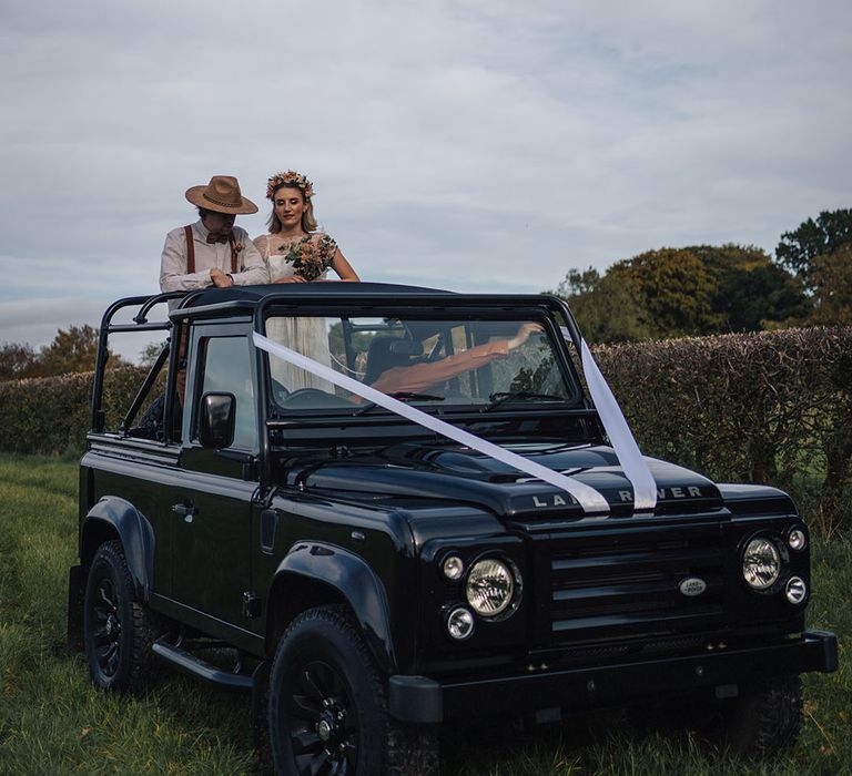 Bride and groom stand in the back of a black Land Rover decorated with white ribbon 