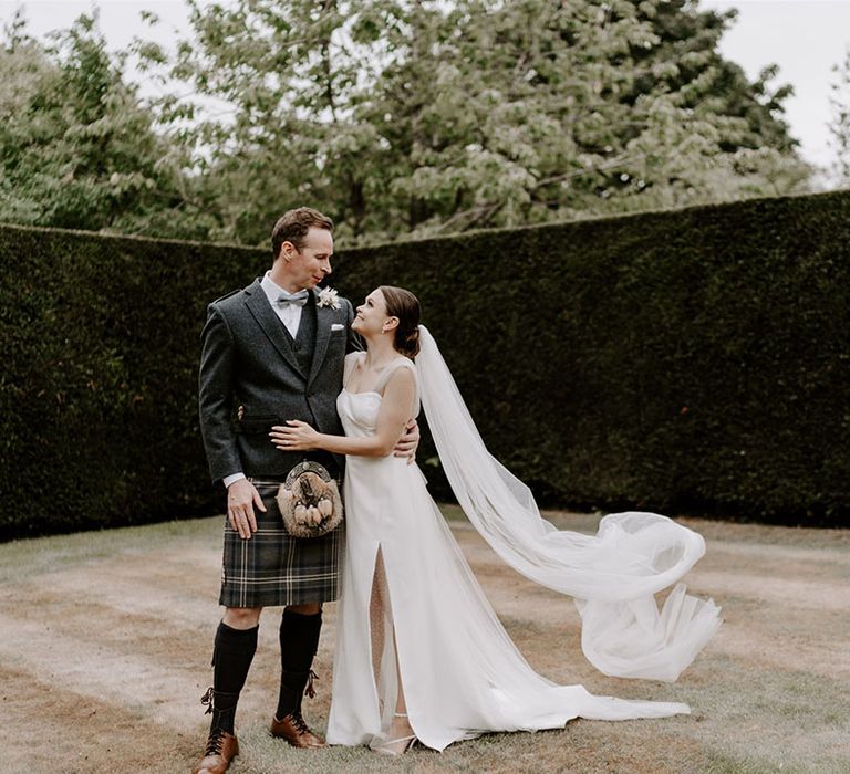 The groom in a kilt and matching tweed waistcoat an d blazer with blue bow tie stands with the bride in a sparkly wedding dress and cathedral length veil 