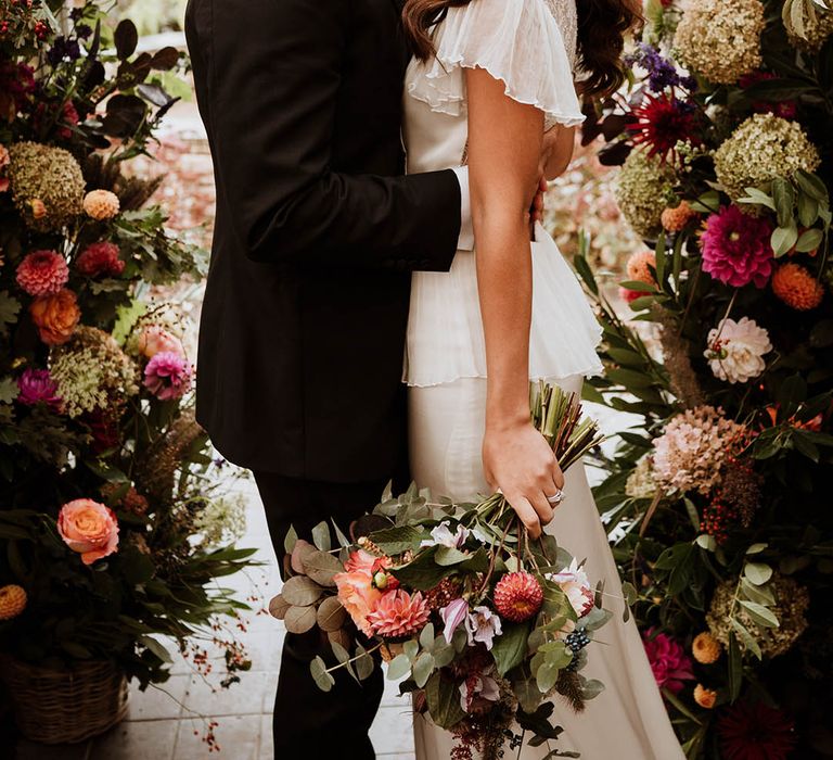 Groom in classic black tuxedo with hand-painted silk cravat and handkerchief kissing bride in Bride in v-neck satin wedding dress with sheer butterfly sleeves and ruffled layer on top standing in large floral arch with baby's-breath, pink and white carnations, garden roses and dried flowers