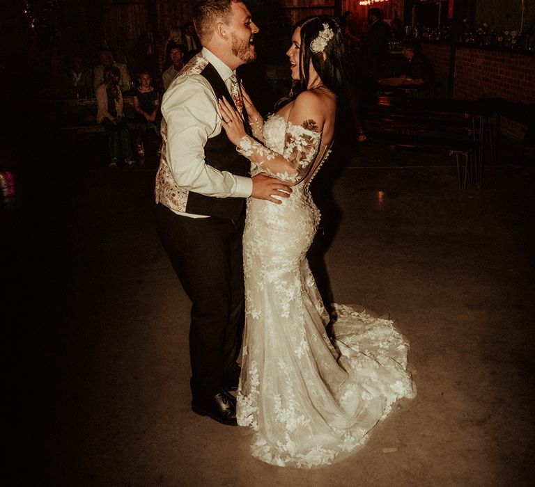 Groom in black waistcoat with patterned back and flower patterned tie and bride in off the shoulder bodycon wedding dress with long, sheer sleeves and lace detailing having their first dance at fun, relaxed weddings