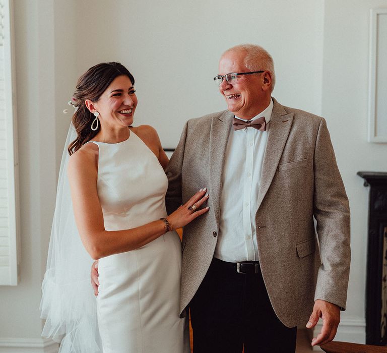 Father of the bride in a beige suit jacket and brown bow tie smiling with the bride in a high neck wedding dress 