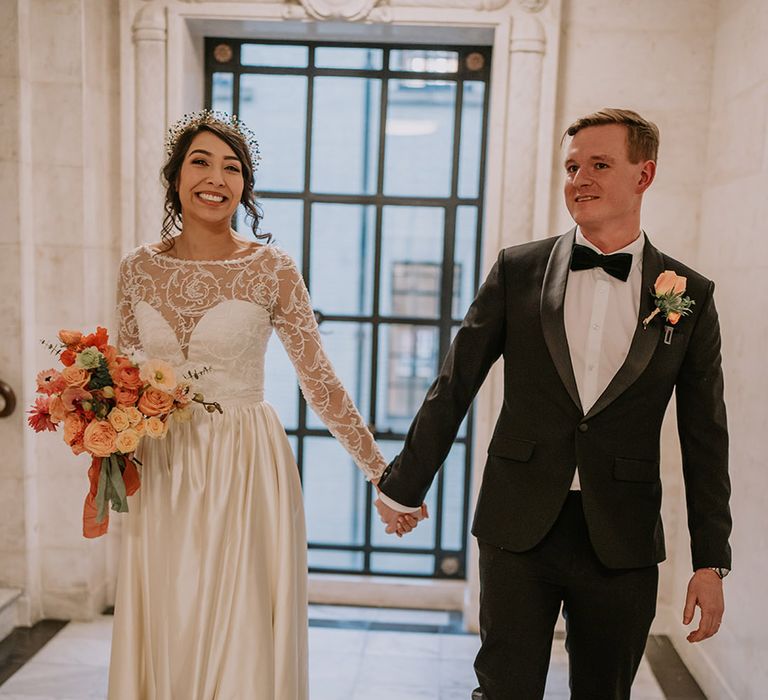 Groom in stylish black tuxedo with black bowtie and light orange rose boutonniere holding hands with bride in  taffeta material princess wedding dress with long lace sleeves 