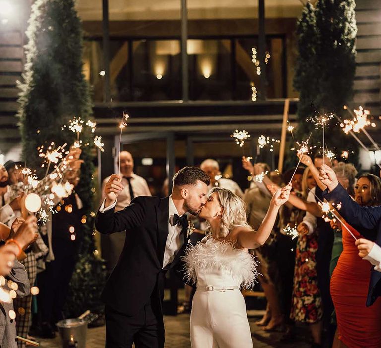 Bride in strapless white satin look feather jumpsuit and groom in classic Ralph Lauren black tuxedo with black bowtie and dried flower boutonniere doing wedding sparkler photoshoot with wedding guests at The Old Kent Barn