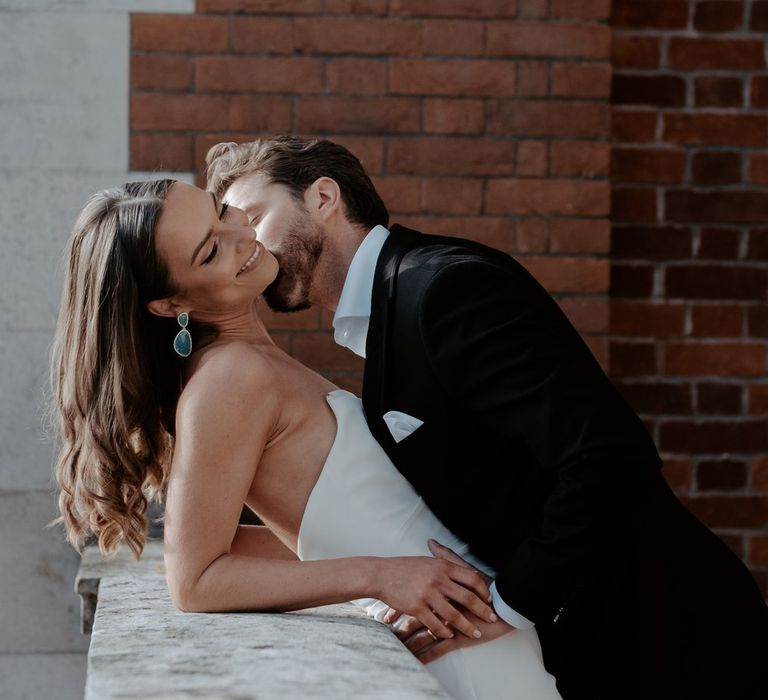 Groom in classic black suit with white pocket square kissing bride in strapless satin wedding dress on the cheek 