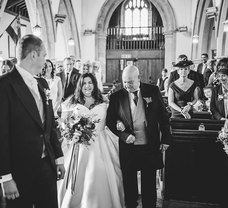 Father of the bride in a morning suit walks the bride down the aisle to the groom in a morning suit for their church wedding 